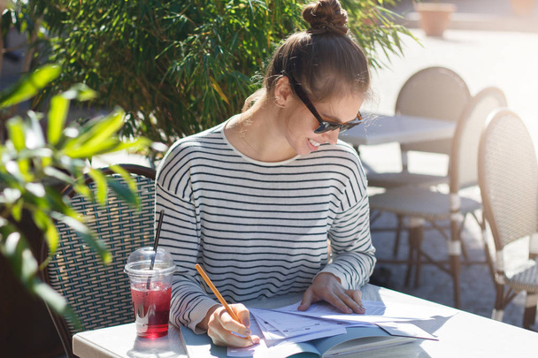 Closeup image of young female wearing trendy sunglasses in street cafe in summer, bending to papers and books lying on table filling important data and checking facts, looking happy and relaxed - Foto, Imagem