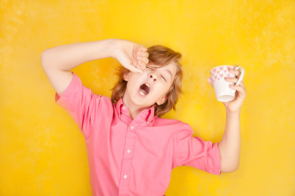 έφηβο φοράει ροζ πουκάμισο στέκεται πάνω σε κίτρινο φόντο στο στούντιο και χασμουρητό με πρωινό καφέ - Φωτογραφία, εικόνα