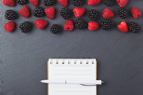 Red raspberries, blackberry, notebook and a pen on the black slate stone background - Photo, Image