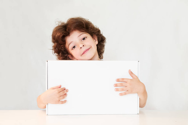 Happy cute boy hugged the white box, smiling and looking at the camera. Gray background. - Foto, imagen