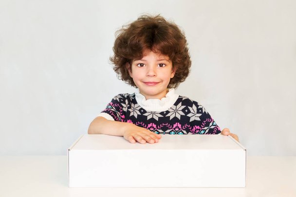 Joyful boy with a cute smile, looking into the camera. He keeps a hand on the white box. Gray background. - Zdjęcie, obraz