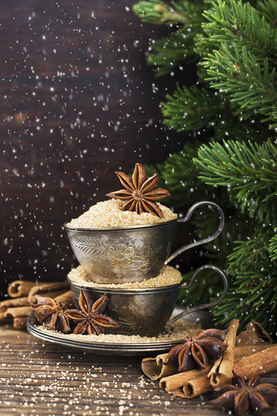 Melchior vintage cups with cane sugar, anise stars, Indian Indian cinnamon sticks on an aged wooden background surrounded by spruce spruce branches. Toned, with imitation of falling snow. Selective fo - Foto, Imagem