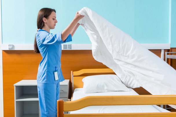 Nuori sairaanhoitaja sinisessä univormussaan vaihtamasta sairaalan sängyn lakanoita
 - Valokuva, kuva