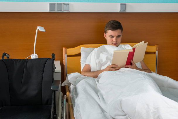 Young male ill patient lying in the hospital bed and reading a b - Valokuva, kuva