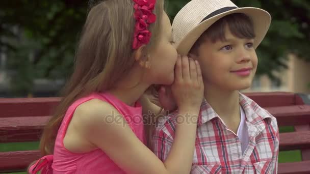 Dziewczynka szepcze do ucha chłopców na ławce - Materiał filmowy, wideo