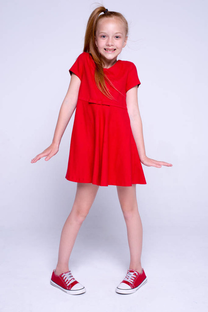 Beautiful little redhead girl in red dress and sneakers posing like model on white background. - Photo, Image
