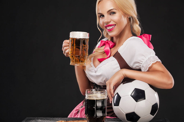 Young sexy Oktoberfest girl - waitress, wearing a traditional Bavarian dress, serving big beer mugs and taking soccer ball on black background. - Photo, Image