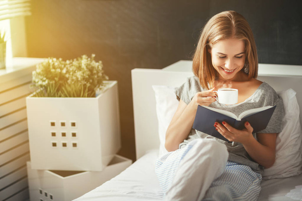Szczęśliwa młoda kobieta czyta książkę i pije kawę w łóżku   - Zdjęcie, obraz