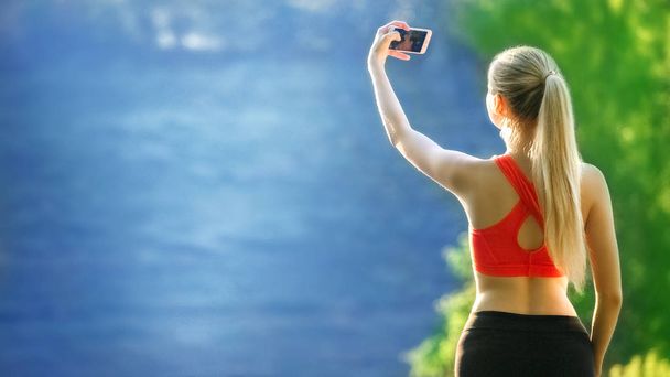 young blonde woman stands on a blue sea background. Sports woman in red t-shirt makes selfie on nature near the sea - Fotoğraf, Görsel