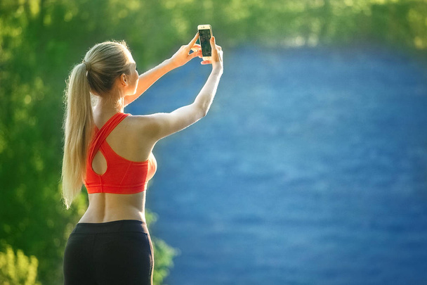 young blonde woman stands on a blue sea background. Sports woman in red t-shirt makes selfie on nature near the sea - Fotoğraf, Görsel