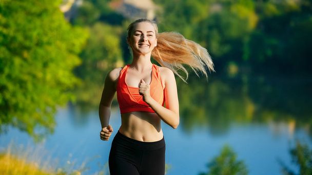 Młoda blond kobieta uruchomiona na charakter. Sportowe dziewczyna w czerwonym T-shirt biegnie wzdłuż parku, w pobliżu rzeki. - Zdjęcie, obraz