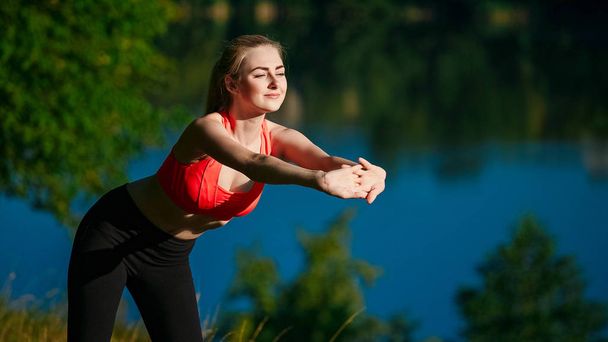 young blond woman doing exercises in nature. A sports girl in a red vest is standing in a park near the river - Foto, afbeelding