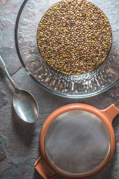 Glass bowl with cannabis seeds, a sieve and a spoon on a stone - Zdjęcie, obraz