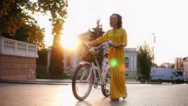Beautiful young woman in a long yellow dress walking during the dawn holding her city bicycles handlebar with flowers in its basket. Lens flare, beautiful city view - Video