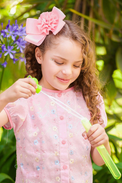 glückliches kleines lockiges Mädchen, das mit Seifenblasen in der sommerlichen Natur spielt, trägt ein rosafarbenes Kleid und eine Blume im Kopf, vor verschwommenem Naturhintergrund - Foto, Bild