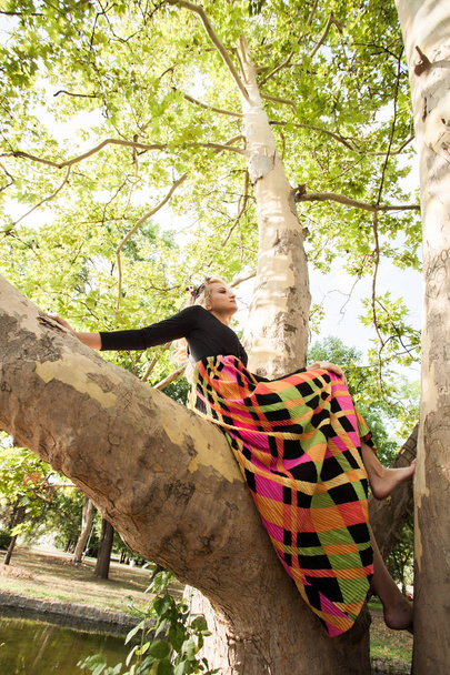 boho girl in long colorful dress sit on tree in park summer day - Photo, Image