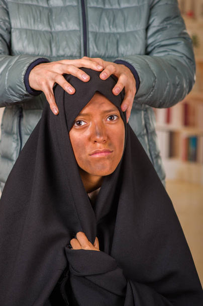 Retrato de uma moça muçulmana vestindo um hijab, com um homem branco atrás dela usando ambas as mãos oprimindo sua cabeça, em um fundo embaçado
 - Foto, Imagem