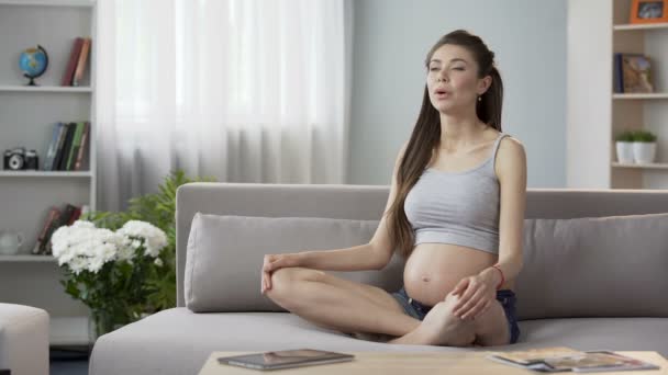 Mother-to-be sitting on couch doing pregnancy yoga breathing exercises, relaxing - Πλάνα, βίντεο