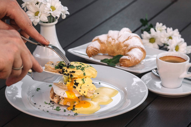 Hands cutting eggs Benedict. Breakfast with poached eggs, croissant, coffee and chamomile on dark wood background - Foto, immagini