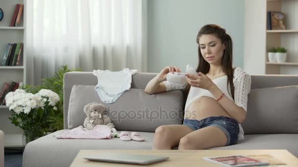 Young pregnant woman taking a look at children clothes on sofa, baby fashion - Πλάνα, βίντεο