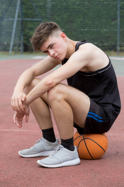 Teenage boy sitting on a basketball on a court looking sad - Fotó, kép