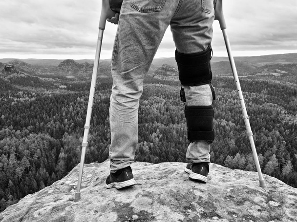 Hombre caminando con muletas en la naturaleza. Hombre piernas en jeans con vendaje ajustable en la rodilla dolor
. - Foto, imagen