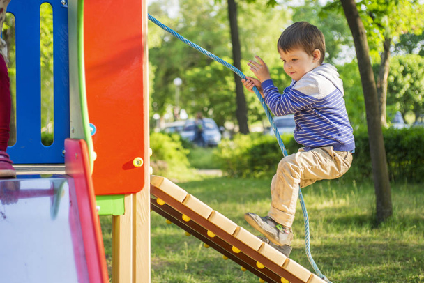 Маленький мальчик взбирается на детский холм, держа верёвку. Ребенок играет на детской площадке
 - Фото, изображение