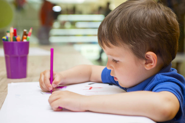 Carino bambino sta disegnando con pennarelli colorati a casa o all'asilo seduto a tavola in una luminosa sala giochi soleggiata
 - Foto, immagini