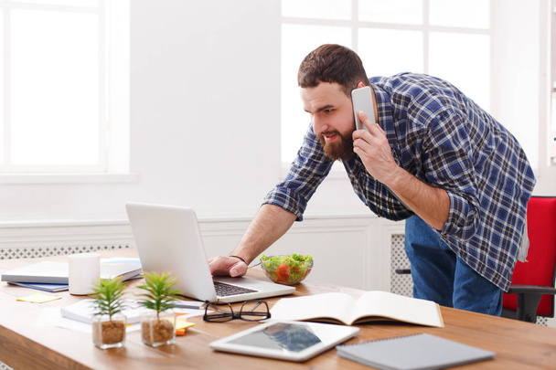 junger Geschäftsmann steht am Laptop und telefoniert in modernem weißen Büro - Foto, Bild