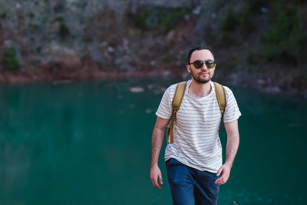 Handsome bearded man, with hands in his pockets,looking away standing near lake. Young guy wearing sunglasses and a fashionable clothes,discover world,stylish cool mans portrait - Φωτογραφία, εικόνα