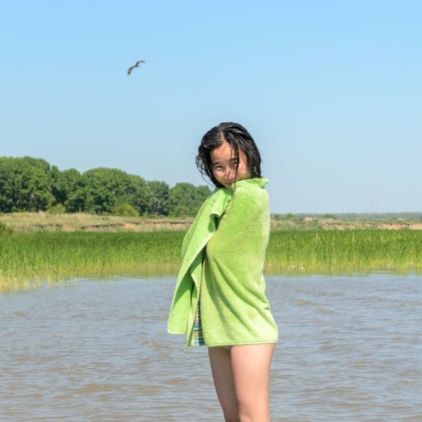 Gyönyörű ázsiai lány mosolyogva van csomagolva egy zöld törülközőt a nedves haj fürdőruhát a háttérben, a madarak, a sas és a fák, az erdő, a szikla és a fű, a víztől. - Fotó, kép