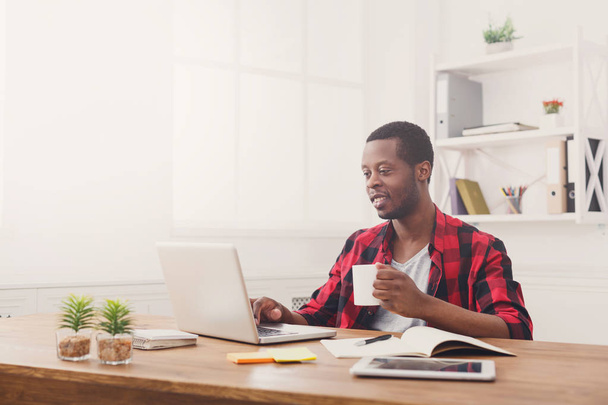 Расслабленный черный бизнесмен в офисе, работает с ноутбуком, пьет кофе
 - Фото, изображение