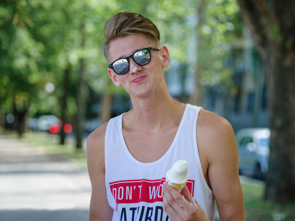 An appealing fellow in sunglasses eating an ice-cream in a park on a natural blurred background. - 写真・画像
