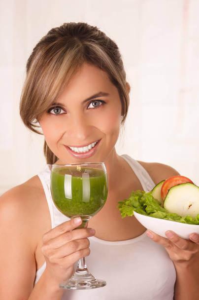 Beautiful young smiling woman wearing a white t-shirt and holding a healthy salad with one hand and glass of juice in her other hand - Фото, зображення