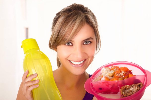 Beautiful smiling woman wearing a purple t-shirt and holding a healthy fruit salad in one hand and a yellow bottle of water in a white background - Photo, image