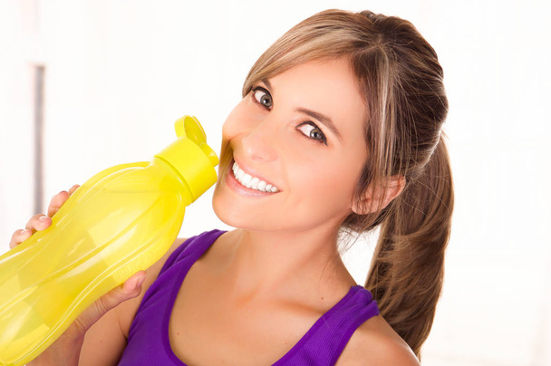 Beautiful smiling woman wearing a purple t-shirt and drinking a yellow bottle of water - Φωτογραφία, εικόνα
