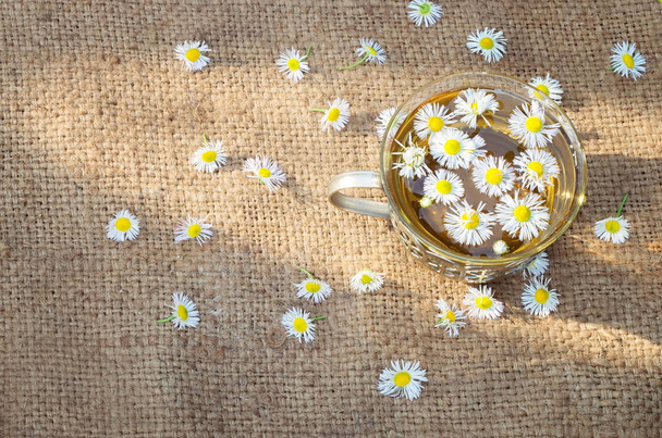 Τσάι χαμομήλι σε φαρδιά ύφασμα, ηλιαχτίδα πρωινού. Σκορπισμένα λουλούδια. - Φωτογραφία, εικόνα