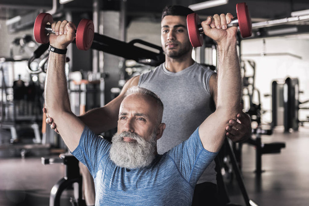 Serious old sportsman doing exercises in modern gym with trainer - Foto, afbeelding