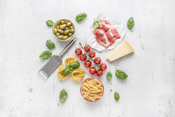 Italian or mediterranean food cuisine and ingredients on white concrete table. Tagliatelle pene pasta  olives olive oil tomatoes parmesan cheese prosciutto and basil leaves on white concrete board - Photo, image
