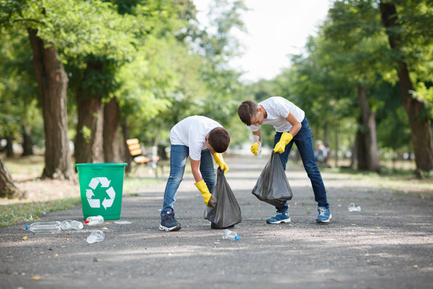 A couple of little boys holding black garbage bags and collecting plastic rubbish on a park background. Ecology protection concept. - 写真・画像