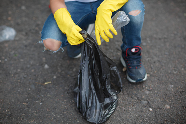 Childs hands in yellow latex gloves holding a black garbage bag on an asphalt background. Ecology protection concept. - Zdjęcie, obraz