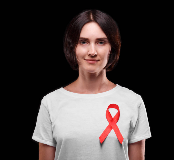 Solidaridad con las personas con SIDA. Una mujer sonriente con una cinta roja en una camiseta sobre un fondo negro. Concepto de concienciación
. - Foto, imagen