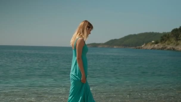 Een vrouw in een lange azuurblauwe jurk wandelingen langs het strand alleen. - Video