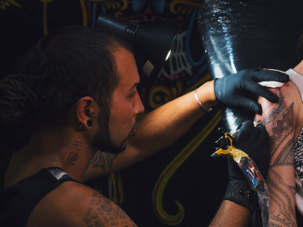 Portrait of man tattoo master with dreadlocks showing process of creation tattoo on female body under the lamp light.Professional artist working in salon, close up view. - Foto, Imagen