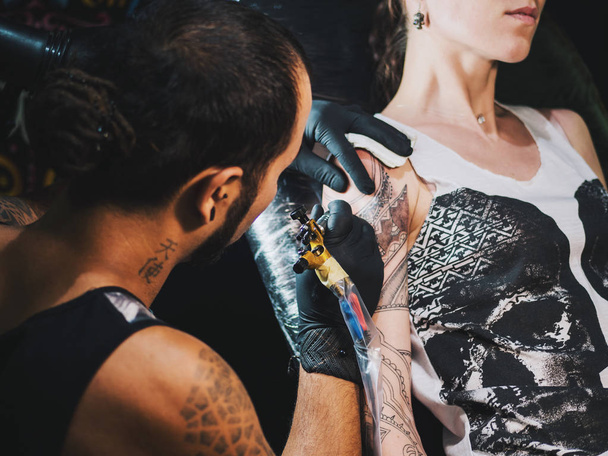 Portrait of man tattoo master with dreadlocks showing process of creation tattoo on female body under the lamp light.Professional artist working in salon, close up view. - Φωτογραφία, εικόνα