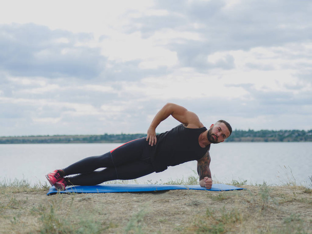 A sporty man with a muscular body doing exercises on a mat on a natural background. Sports and yoga concept. - Foto, Bild