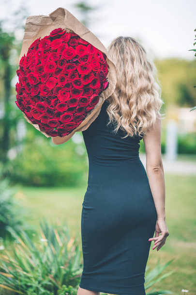 Back view of elegant beautiful woman is wearing black fashion dress is holding big bouquet of 101 red roses - Φωτογραφία, εικόνα