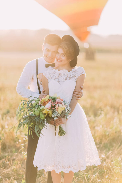 Smiling groom is hugging the vintage dressed bride with the huge bouquet back at the background of the sunny field. - Foto, Imagem