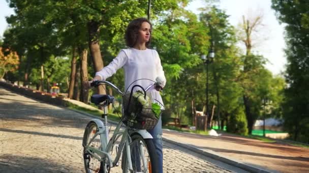 Young woman in a white t-shirt and blue jeans walking on the cobblestone road in the city park holding her city bicycles handlebar with flowers in its basket. Slowmotion shot - Filmati, video