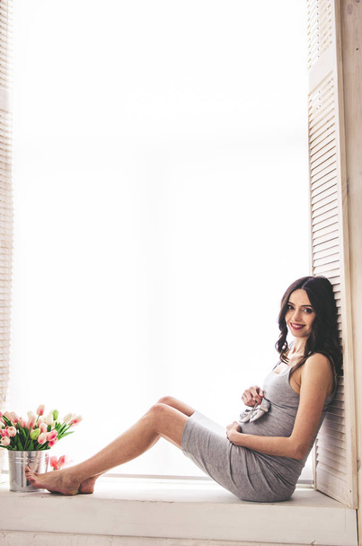 Schwangere Frau in grauem Kleid sitzt am Fenster  - Foto, Bild